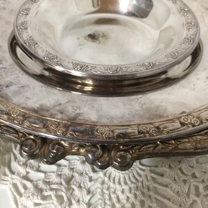 Puede incluir: Un plato de servir de plata con un borde elevado y dise&ntilde;os florales intrincados. El plato tiene un taz&oacute;n m&aacute;s peque&ntilde;o y extra&iacute;ble en el centro.