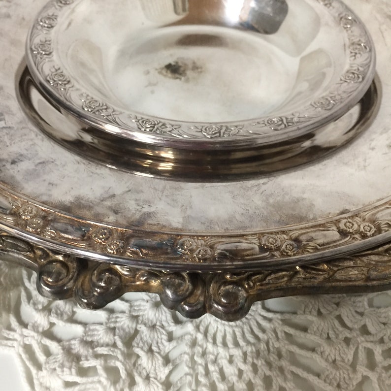 Puede incluir: Un plato de servir de plata con un borde elevado y dise&ntilde;os florales intrincados. El plato tiene un taz&oacute;n m&aacute;s peque&ntilde;o y extra&iacute;ble en el centro.