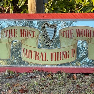 K&ouml;nnte beinhalten: Ein roter Spiegelrahmen mit einem goldenen und wei&szlig;en Design. Der Spiegel tr&auml;gt den Text "The Most Natural Thing In The World" mit einer Harfe in der Mitte.