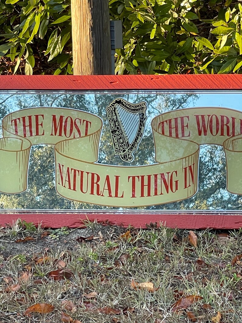 K&ouml;nnte beinhalten: Ein roter Spiegelrahmen mit einem goldenen und wei&szlig;en Design. Der Spiegel tr&auml;gt den Text "The Most Natural Thing In The World" mit einer Harfe in der Mitte.