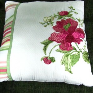 May include: White pillow with a green and pink striped border. The pillow features an embroidered floral design with pink flowers and green leaves.