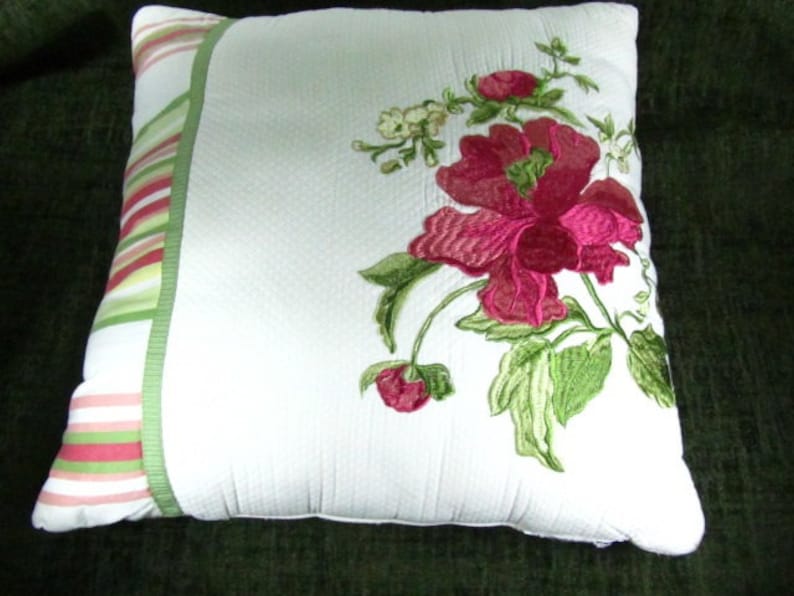 May include: White pillow with a green and pink striped border. The pillow features an embroidered floral design with pink flowers and green leaves.