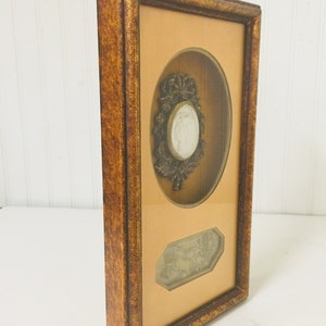 May include: A framed artwork featuring a raised oval cameo with a woman's profile and a floral design. The frame is made of wood with a dark brown finish. The artwork is set against a light brown background.