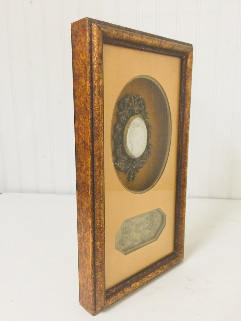 May include: A framed artwork featuring a raised oval cameo with a woman's profile and a floral design. The frame is made of wood with a dark brown finish. The artwork is set against a light brown background.