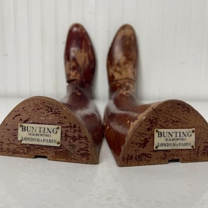 May include: A pair of wooden shoe trees with a triangular base. The shoe trees are brown and have a label that reads "Bunting (J.R. Bunting) London & Paris".