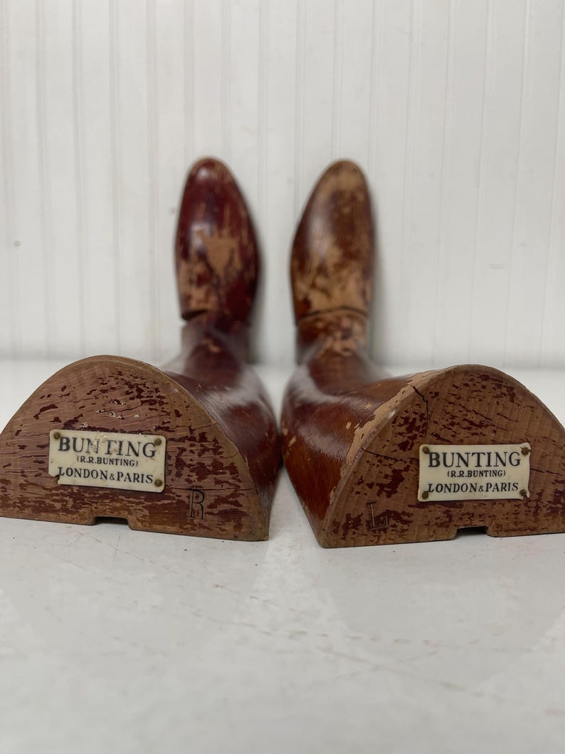 May include: A pair of wooden shoe trees with a triangular base. The shoe trees are brown and have a label that reads "Bunting (J.R. Bunting) London & Paris".