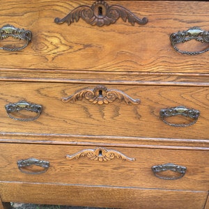 May include: A wooden dresser with three drawers and ornate metal handles. The dresser is a light brown color and has a distressed finish.