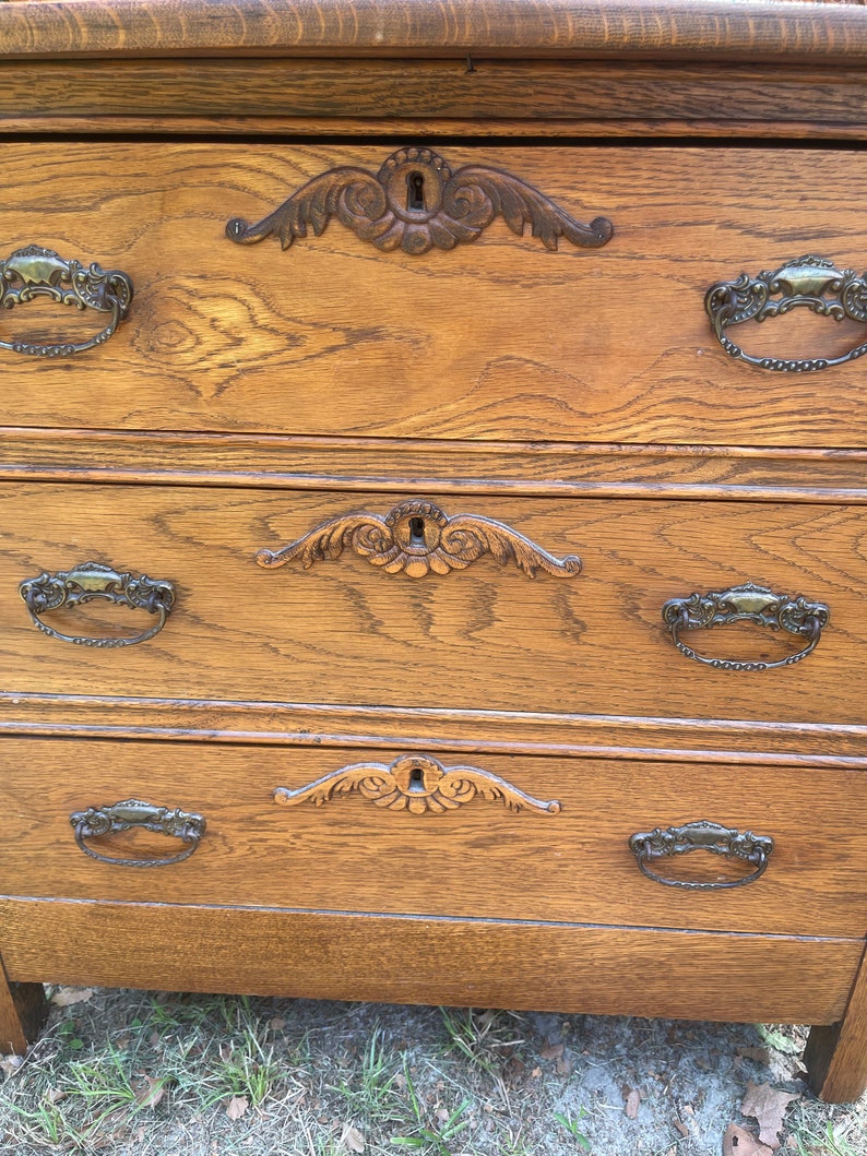 May include: A wooden dresser with three drawers and ornate metal handles. The dresser is a light brown color and has a distressed finish.