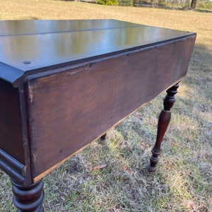 May include: A dark brown wooden desk with turned legs. The desk has a drawer on the left side and a shelf on the right side. The desk is sitting on a grassy lawn.