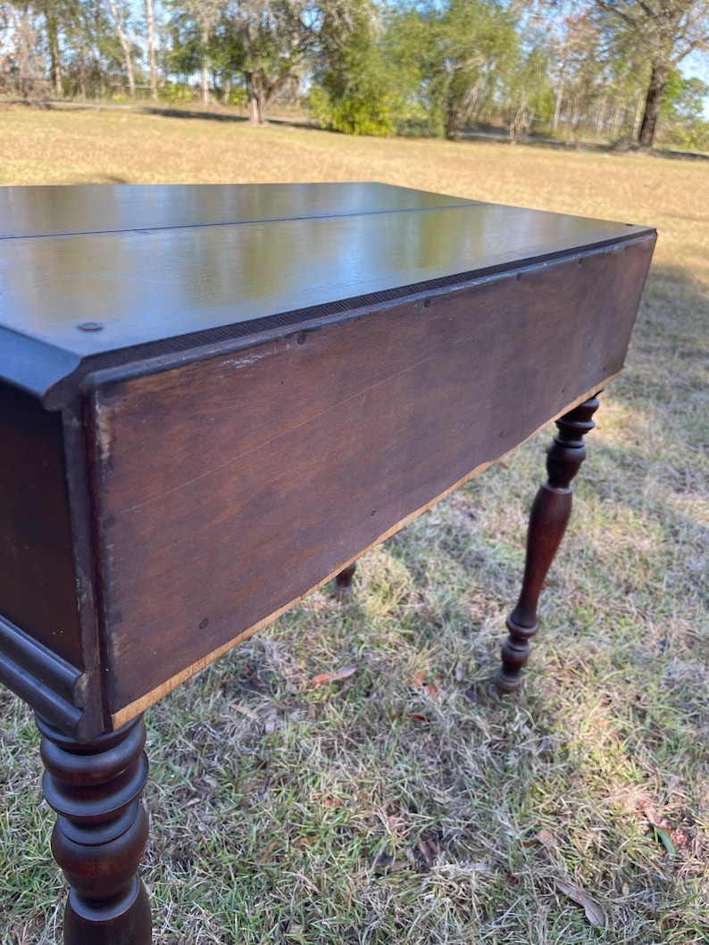 May include: A dark brown wooden desk with turned legs. The desk has a drawer on the left side and a shelf on the right side. The desk is sitting on a grassy lawn.