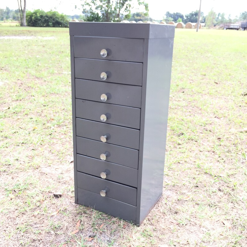 May include: A gray metal drawer cabinet with eight drawers and silver knobs. The cabinet is standing on a grassy surface.