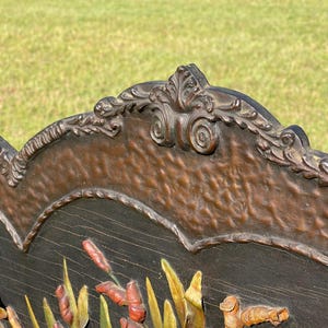 May include: Close-up of a wooden decorative panel with a carved design. The panel features a dark brown background with a relief of cattails and other marsh plants in shades of red, green, and yellow. The top edge has an ornate, scrolled border.
