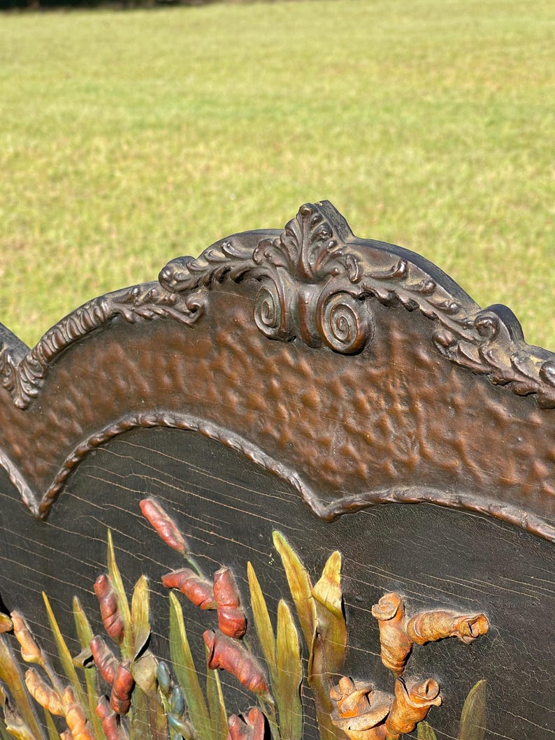 May include: Close-up of a wooden decorative panel with a carved design. The panel features a dark brown background with a relief of cattails and other marsh plants in shades of red, green, and yellow. The top edge has an ornate, scrolled border.