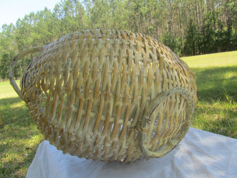 May include: A large, whitewashed wicker basket with a woven handle. The basket is made of natural materials and has a rustic, vintage look.