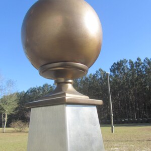 May include: A gold metal finial with a round top and a square base. The finial is mounted on a silver metal post.