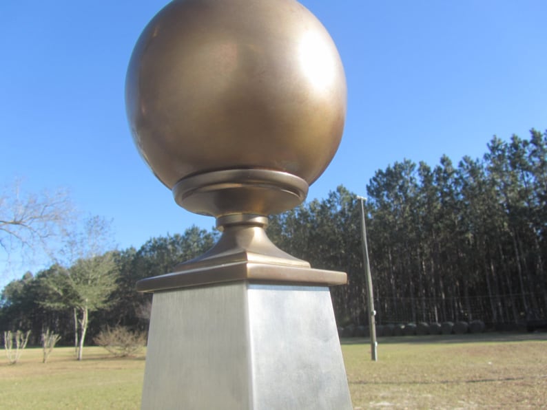 May include: A gold metal finial with a round top and a square base. The finial is mounted on a silver metal post.