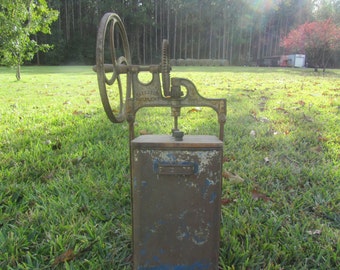 May include: A vintage, rusty, metal apple cider press with a hand crank and a metal container. The press is labeled "Waterloo Excelsior" and is sitting on a grassy surface.
