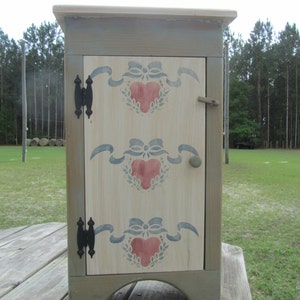 May include: A small, wooden cabinet with a light gray paint finish and a white door. The door has a painted design of blue ribbons and red hearts. The cabinet has two black hinges and a small knob.