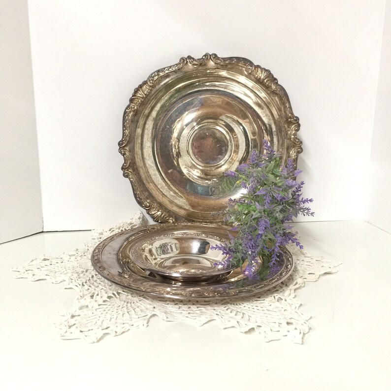 Puede incluir: Tres platos de servir plateados, uno grande y dos m&aacute;s peque&ntilde;os, est&aacute;n apilados sobre un encaje de ganchillo blanco. Los platos tienen un borde decorativo y adornado. Un ramo de flores moradas est&aacute; delante de los platos.