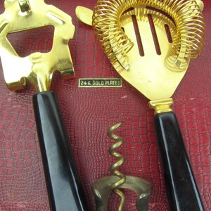 Op de afbeelding: Een set van drie vergulde bargereedschappen met zwarte kunststof handvatten. De gereedschappen omvatten een flessenopener, een cocktailzeef en een kurkentrekker. De gereedschappen zijn gemarkeerd met "24 K. GOLD PLATED".