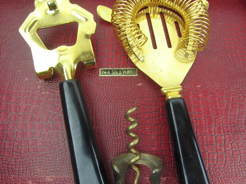 Op de afbeelding: Een set van drie vergulde bargereedschappen met zwarte kunststof handvatten. De gereedschappen omvatten een flessenopener, een cocktailzeef en een kurkentrekker. De gereedschappen zijn gemarkeerd met "24 K. GOLD PLATED".