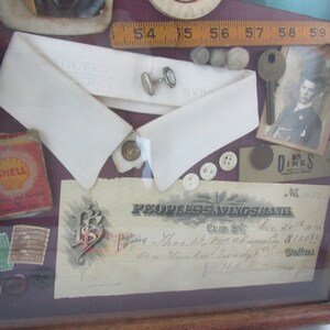 Puede incluir: Una caja de sombra enmarcada con una variedad de art&iacute;culos, que incluyen un cuello blanco, una peque&ntilde;a regla de madera, una llave, una peque&ntilde;a imagen de una persona, un peque&ntilde;o trozo de papel con el texto "$5 Dimes", un peque&ntilde;o trozo de papel con el texto "PEOPLES SAVINGS BANK", un peque&ntilde;o trozo de papel con el texto "CLIO S.C.", un peque&ntilde;o trozo de papel con el texto "Dec 20th 1900", un peque&ntilde;o trozo de papel con el texto "$12.50", un peque&ntilde;o trozo de papel con el texto "Dollars", un peque&ntilde;o trozo de papel con el texto "N.L. 377", un peque&ntilde;o trozo de papel con el texto "SHELL", algunos botones y algunos otros art&iacute;culos peque&ntilde;os.