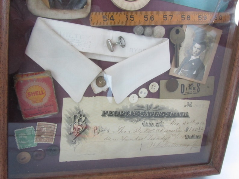 Puede incluir: Una caja de sombra enmarcada con una variedad de art&iacute;culos, que incluyen un cuello blanco, una peque&ntilde;a regla de madera, una llave, una peque&ntilde;a imagen de una persona, un peque&ntilde;o trozo de papel con el texto "$5 Dimes", un peque&ntilde;o trozo de papel con el texto "PEOPLES SAVINGS BANK", un peque&ntilde;o trozo de papel con el texto "CLIO S.C.", un peque&ntilde;o trozo de papel con el texto "Dec 20th 1900", un peque&ntilde;o trozo de papel con el texto "$12.50", un peque&ntilde;o trozo de papel con el texto "Dollars", un peque&ntilde;o trozo de papel con el texto "N.L. 377", un peque&ntilde;o trozo de papel con el texto "SHELL", algunos botones y algunos otros art&iacute;culos peque&ntilde;os.