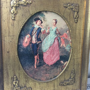 May include: A framed oval painting of a couple in a garden setting. The man is wearing a blue jacket and the woman is wearing a pink dress. The painting is set in a gold frame with ornate corners.
