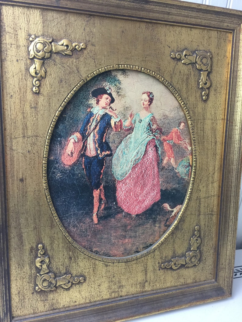 May include: A framed oval painting of a couple in a garden setting. The man is wearing a blue jacket and the woman is wearing a pink dress. The painting is set in a gold frame with ornate corners.