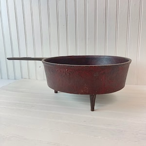 May include: A red cast iron pot with three legs and a handle. The pot is sitting on a white wooden surface.