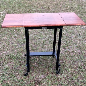 May include: A wooden table with a black metal frame and wheels. The table has a drop leaf on each side. The table is labeled "The Folding Table Co."