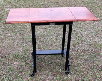 Puede incluir: Una mesa de madera con un marco de metal negro y ruedas. La mesa tiene una hoja abatible a cada lado. La mesa está etiquetada "The Folding Table Co."