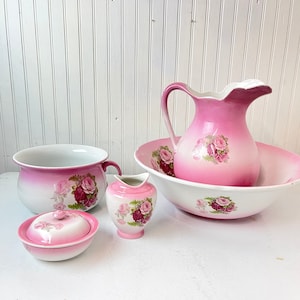 May include: A set of five pink and white porcelain bathroom accessories with a floral design. The set includes a pitcher, a bowl, a lidded jar, and two small vases.