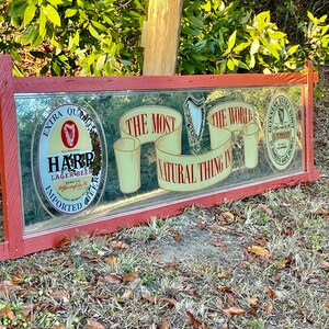K&ouml;nnte beinhalten: Ein roter Spiegelrahmen mit einer Harp Lager Beer-Werbung. Die Anzeige zeigt eine Harfe, ein Glas Bier und den Text "Das nat&uuml;rlichste der Welt".