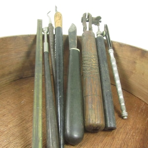 May include: A collection of antique tools, including various metal and wooden handled instruments. The tools are arranged inside a wooden bowl, showcasing a variety of shapes and sizes, with some appearing to be for carving or fine detail work.
