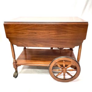 May include: A vintage wooden tea cart with a rectangular top and a lower shelf. The cart features decorative spoked wheels and is made of a rich, warm-toned wood. The cart has a classic design with a polished finish.
