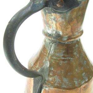 May include: An antique copper pitcher with a hammered finish and a worn, dark brown iron handle. The pitcher has a wide, round body and a narrow neck.