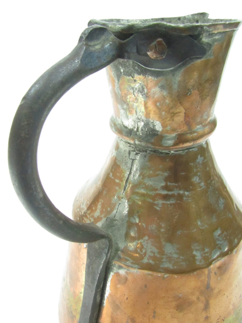 May include: An antique copper pitcher with a hammered finish and a worn, dark brown iron handle. The pitcher has a wide, round body and a narrow neck.