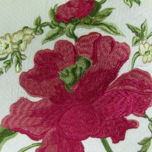 May include: Embroidered floral design featuring a large pink flower with green leaves and smaller white flowers. The design is on a white background.