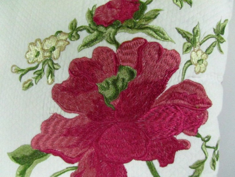May include: Embroidered floral design featuring a large pink flower with green leaves and smaller white flowers. The design is on a white background.