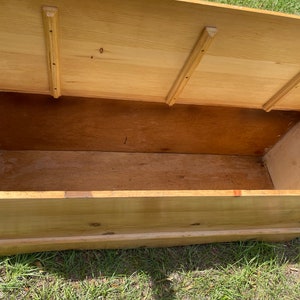 K&ouml;nnte beinhalten: Eine Holzkiste mit einem Scharnierdeckel, ge&ouml;ffnet, um den leeren Innenraum zu zeigen. Die Kiste ist aus hellem Holz gefertigt und hat ein schlichtes Design.