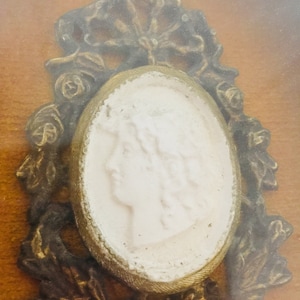 May include: A white cameo portrait of a woman with a gold frame surrounded by ornate gold floral details.