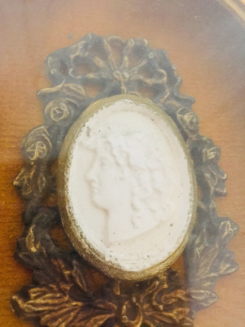 May include: A white cameo portrait of a woman with a gold frame surrounded by ornate gold floral details.