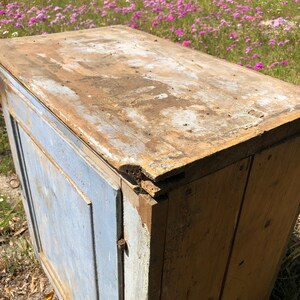 May include: A vintage wooden cabinet with a single door and a top shelf. The cabinet is painted a light blue color and has a distressed finish. The top shelf is made of natural wood and has a weathered look.