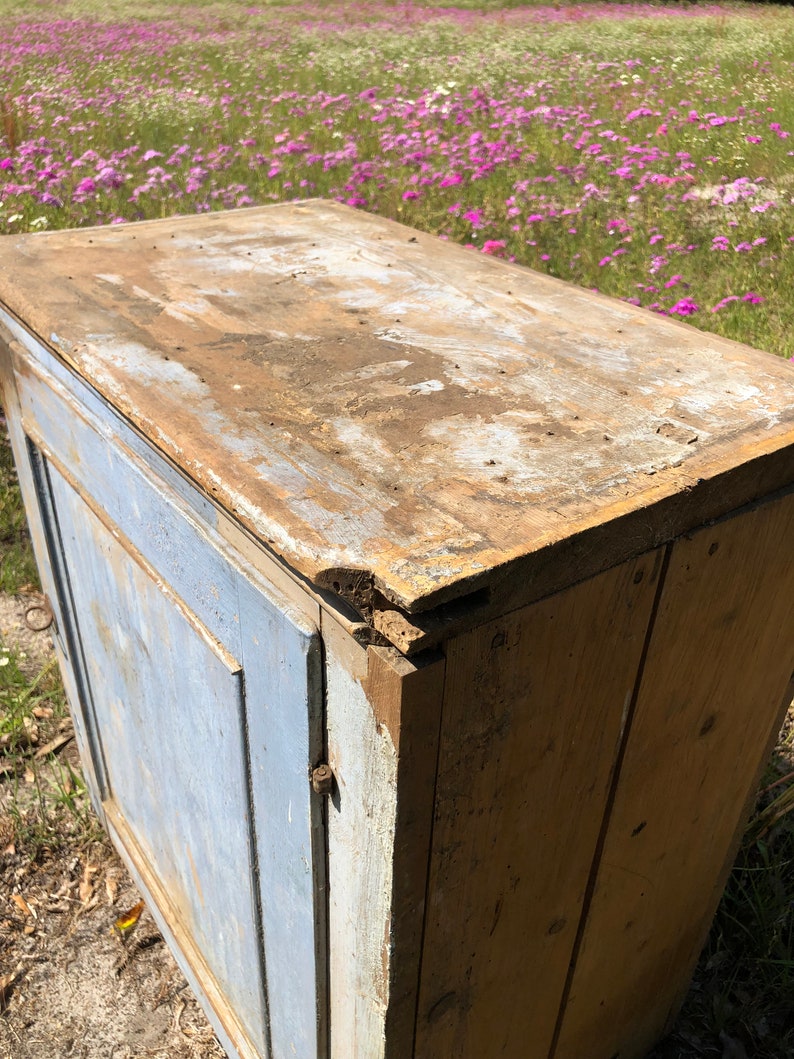 May include: A vintage wooden cabinet with a single door and a top shelf. The cabinet is painted a light blue color and has a distressed finish. The top shelf is made of natural wood and has a weathered look.