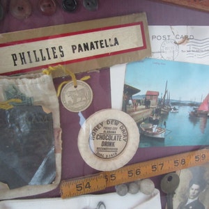 Puede incluir: Un collage enmarcado de objetos vintage que incluye una peque&ntilde;a bolsa, una postal, una regla, una peque&ntilde;a ficha de metal, una peque&ntilde;a etiqueta de metal y un peque&ntilde;o disco de metal. La postal presenta una foto de un puerto con barcos y una vela roja. La ficha de metal tiene el texto "Honey Dew Dairy" y "Chocolate Drink". La etiqueta de metal tiene el texto "Phillies Panatella".