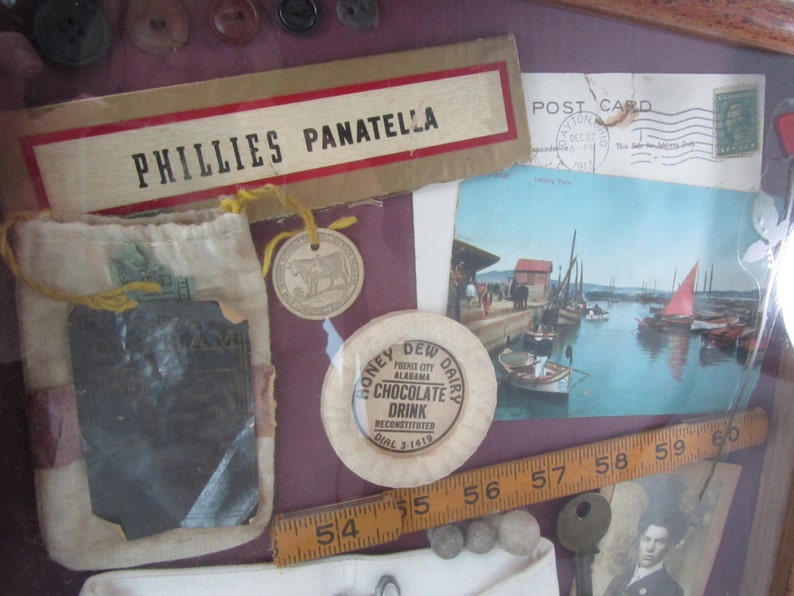 Puede incluir: Un collage enmarcado de objetos vintage que incluye una peque&ntilde;a bolsa, una postal, una regla, una peque&ntilde;a ficha de metal, una peque&ntilde;a etiqueta de metal y un peque&ntilde;o disco de metal. La postal presenta una foto de un puerto con barcos y una vela roja. La ficha de metal tiene el texto "Honey Dew Dairy" y "Chocolate Drink". La etiqueta de metal tiene el texto "Phillies Panatella".