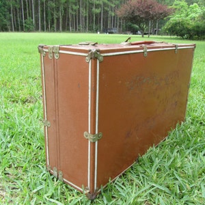 May include: A vintage brown suitcase with brass hardware and white trim. The suitcase is lying on its side on a grassy surface.