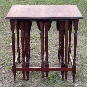 May include: A set of four nesting tables made of dark wood with a rectangular top and turned legs. The tables are stacked on top of each other, with the largest table at the bottom and the smallest table at the top.