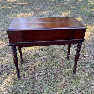 May include: A dark brown wooden writing desk with a drop-down front and turned legs. The desk is sitting on a green grassy surface.